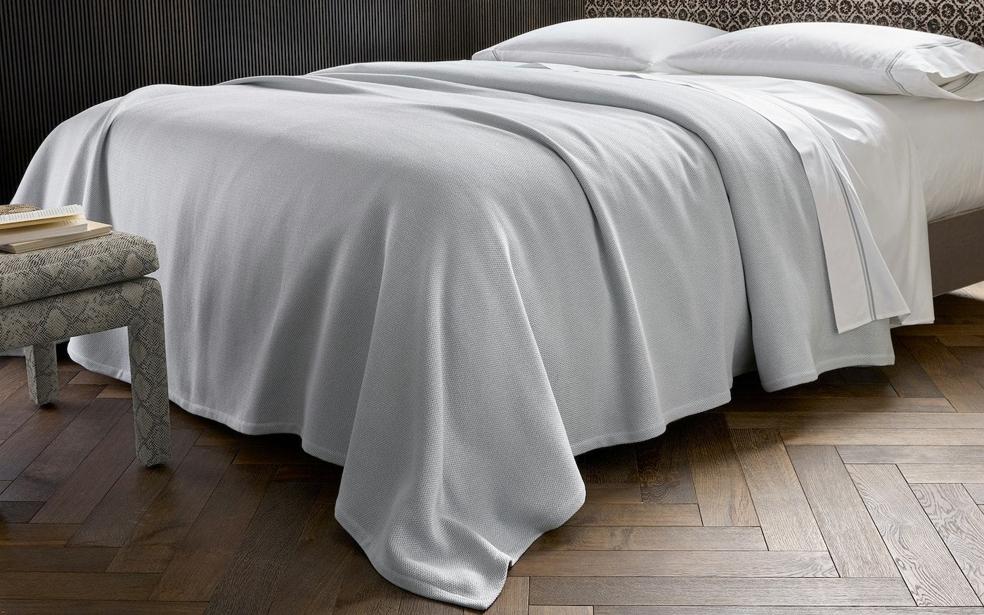 A neatly made bed with white bedding is accented by the soft, knitted Costa Mesa Blanket Collection from Matouk, draped beside a patterned bench on a wooden herringbone floor.
