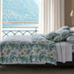 A neatly made bed showcases the Matouk Del Tesoro Bedding Collection in Sea, set by a window with lake and mountain views, complemented by curtains and a nightstand.