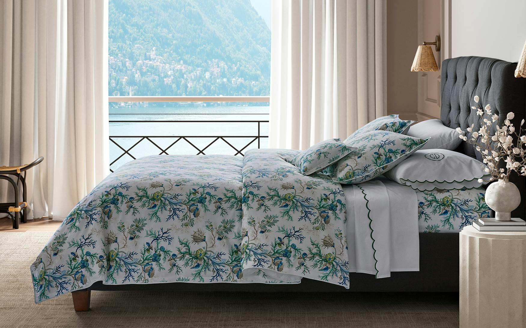 A neatly made bed showcases the Matouk Del Tesoro Bedding Collection in Sea, set by a window with lake and mountain views, complemented by curtains and a nightstand.
