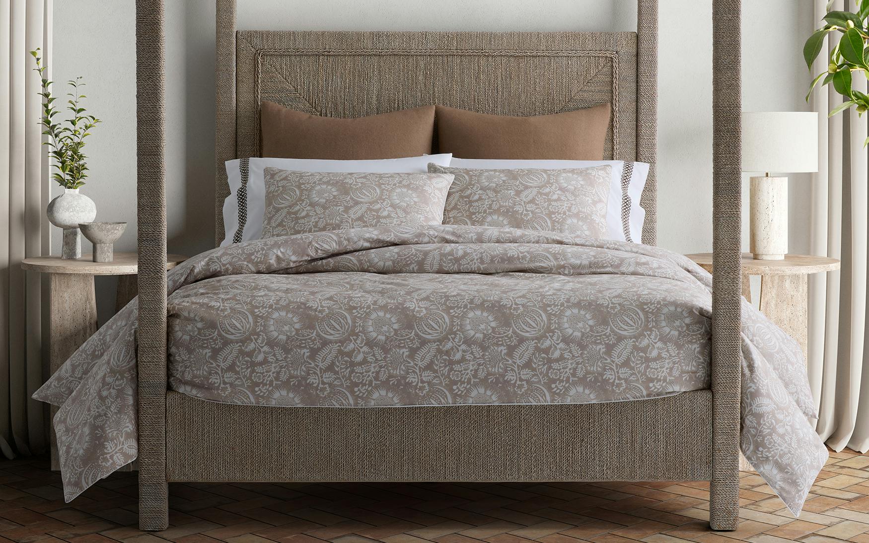 A neatly made bed features the Matouk Granada Bedding Collection in Dune, with a patterned beige cotton percale comforter and matching pillows, set between two small round bedside tables adorned with plants and decor.