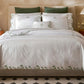 A neatly made bed features the Matouk Marcella Bedding Collection in Clover—white embroidered bedding with scalloped florals—three pillows, and two bedside tables with lamps, all set in a room with patterned floor tiles.