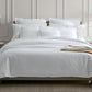 The bed is adorned with the luxurious Matouk White Roman Hemstitch Bedding Collection in a well-lit room with beige walls. White sheets, pillows, and blankets complement two wall-mounted lamps and a side table with a glass.