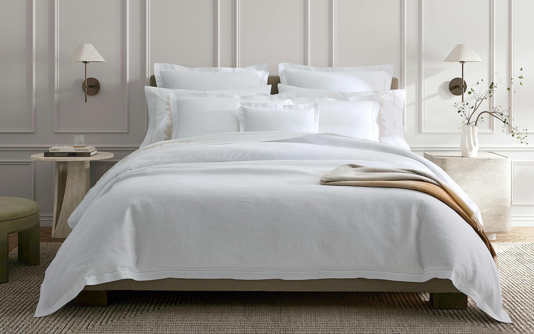 The bed is adorned with the luxurious Matouk White Roman Hemstitch Bedding Collection in a well-lit room with beige walls. White sheets, pillows, and blankets complement two wall-mounted lamps and a side table with a glass.