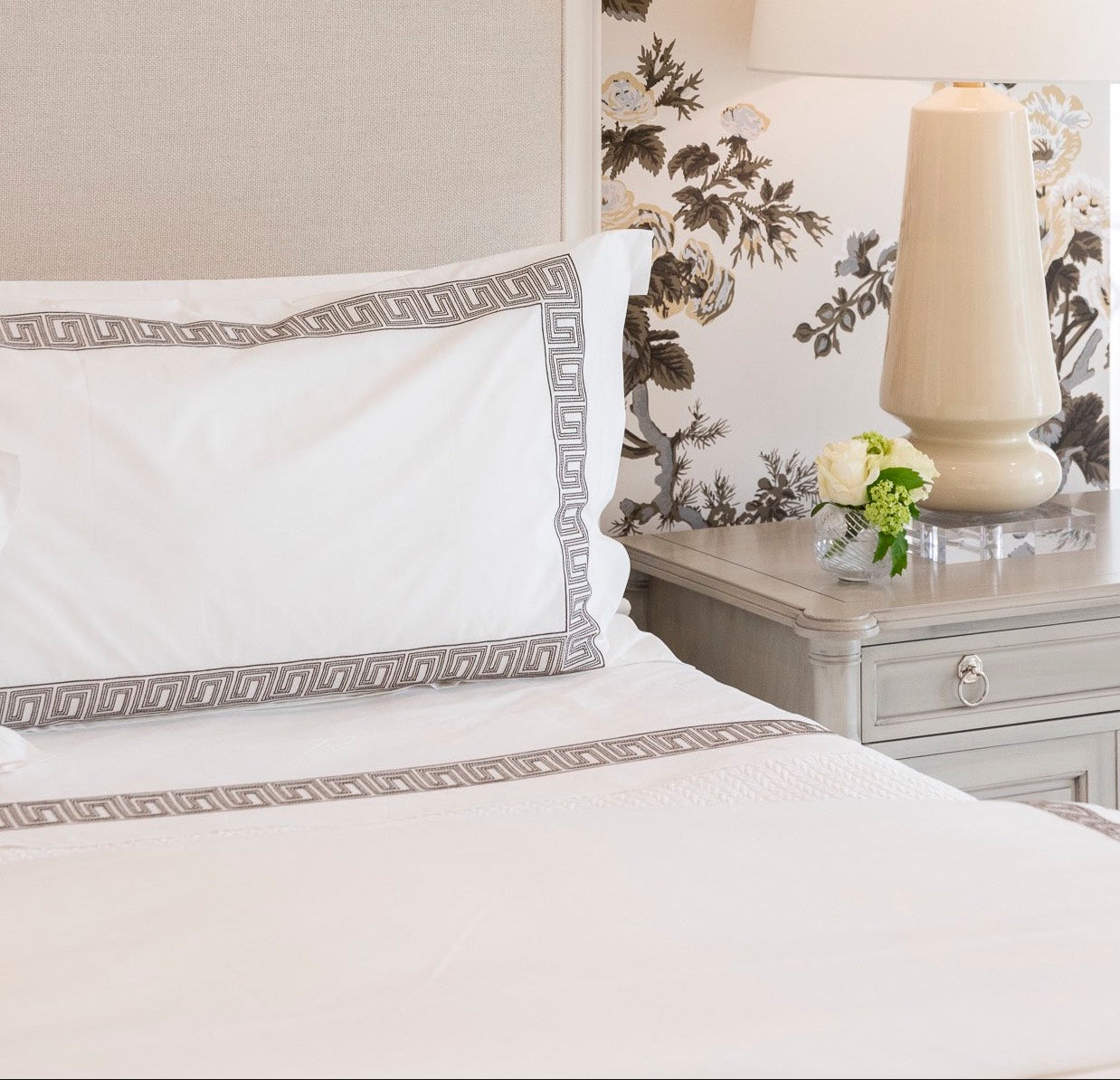 A neatly made bed with the Matouk Ophelia Bedding Collection in Mocha sits beside a gray nightstand topped with a beige lamp and a small vase of white flowers.