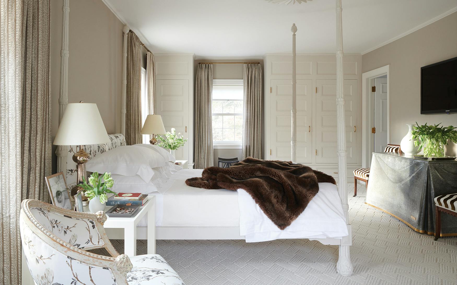 A neatly made bed with Matouk's Luca 14" Fitted Sheet Collection in white and a dark brown throw sits in a bright bedroom featuring neutral walls, patterned curtains, and modern furniture.