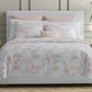 A neatly made bed with Matouk's Dominique Bedding Collection in Blush, crafted from OEKO-TEX® certified Egyptian cotton percale, flanked by two side tables with lamps, a plant, and neutral-colored walls in the background.