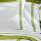 Matouk’s Astor Braid Bedding Collection in Grass, featuring white linens with a green embroidered border, is displayed on a bed with green accents in the background.