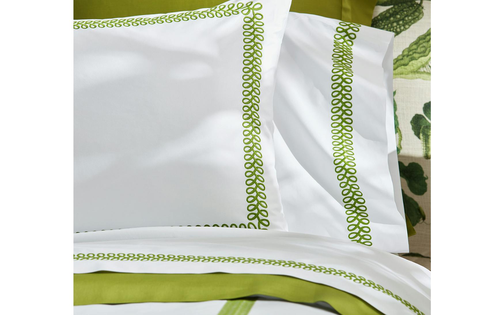 Matouk’s Astor Braid Bedding Collection in Grass, featuring white linens with a green embroidered border, is displayed on a bed with green accents in the background.
