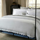 A neatly made bed with Matouk’s Butterfield Bedding Collection in Ocean, featuring scallop details and multiple pillows, set in a room with large windows, beige curtains, and a dark dresser in the background.