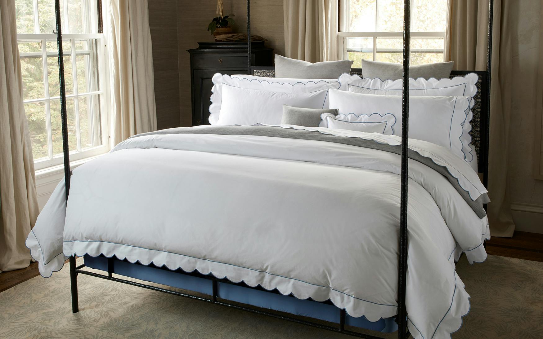 A neatly made bed with Matouk’s Butterfield Bedding Collection in Ocean, featuring scallop details and multiple pillows, set in a room with large windows, beige curtains, and a dark dresser in the background.