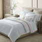 A neatly made bed adorned with the luxurious Matouk Callista Bedding Collection in white and beige Giza Egyptian cotton, featuring 500 thread count sheets and decorative embroidery on the pillows, is complemented by a matching nightstand and dresser in a modern, softly lit bedroom.