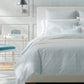 A neatly made bed with Matouk's Astor Braid Bedding Collection in Sky, a headboard, a white side table with a blue box, and a wall-mounted silver lamp.