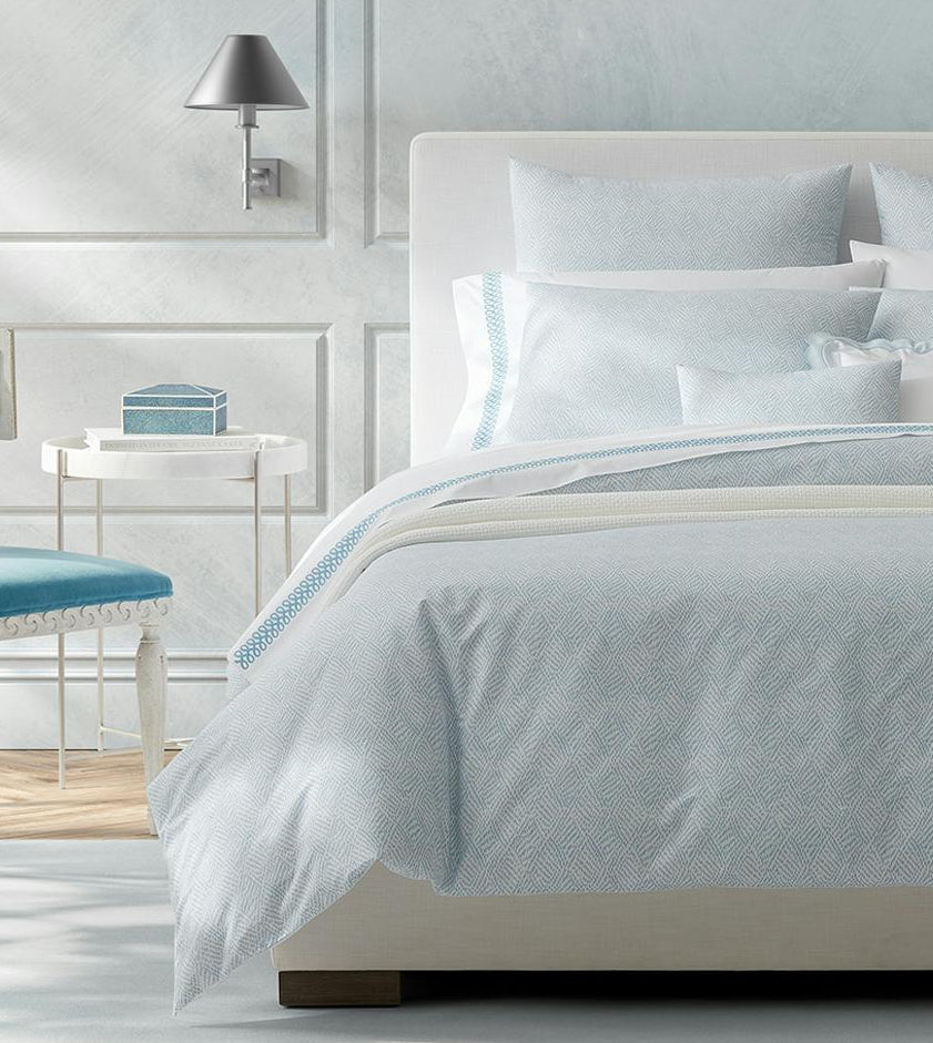 A neatly made bed with Matouk's Astor Braid Bedding Collection in Sky, a headboard, a white side table with a blue box, and a wall-mounted silver lamp.