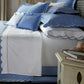 A neatly made bed is styled with the Matouk Butterfield Bedding Collection, featuring blue and white linens adorned with elegant scalloped edges, complemented by pillows showcasing delicate scallop detailing; beside the bed, a nightstand displays books, jewelry, and a decorative accent.