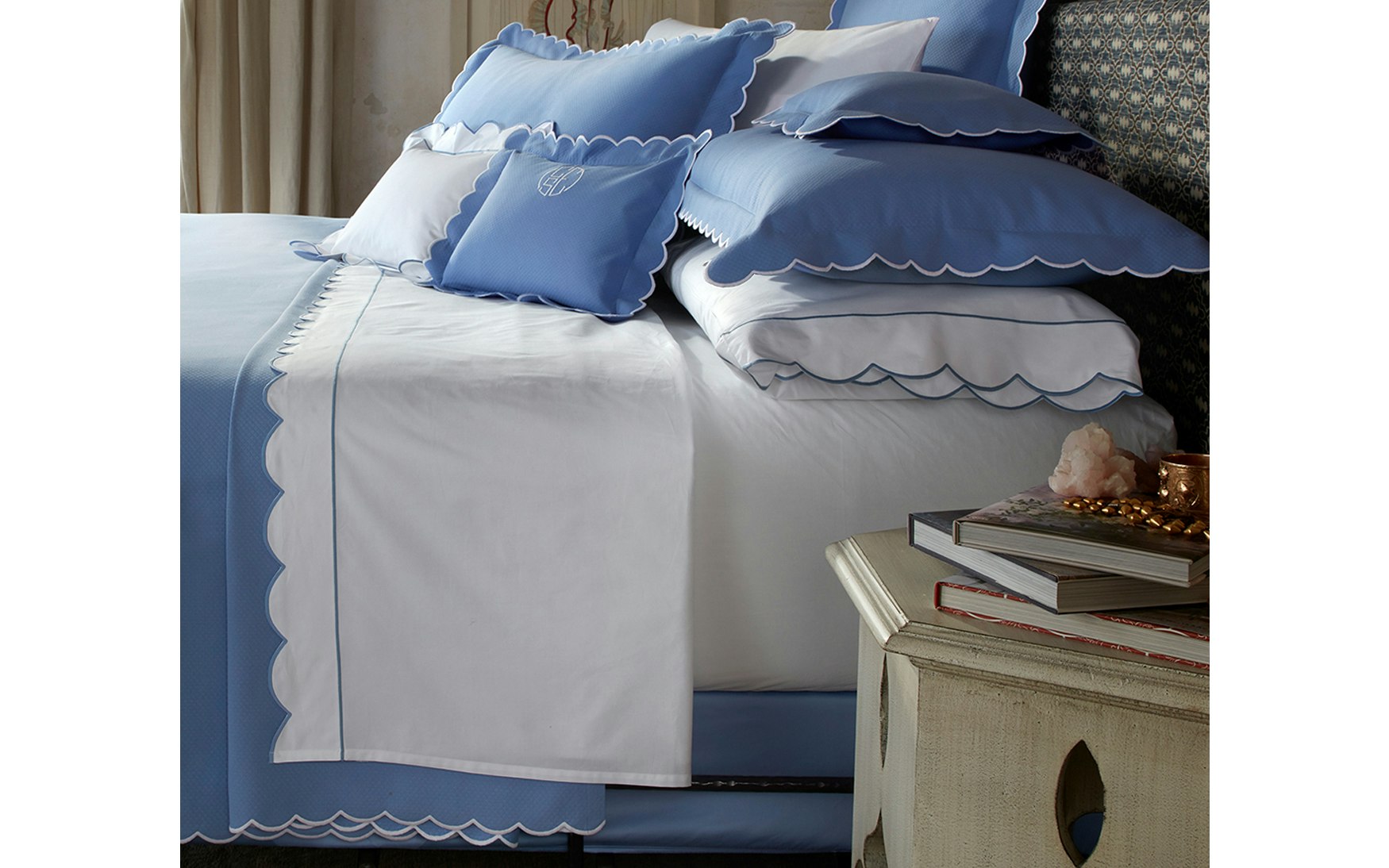 A neatly made bed is styled with the Matouk Butterfield Bedding Collection, featuring blue and white linens adorned with elegant scalloped edges, complemented by pillows showcasing delicate scallop detailing; beside the bed, a nightstand displays books, jewelry, and a decorative accent.