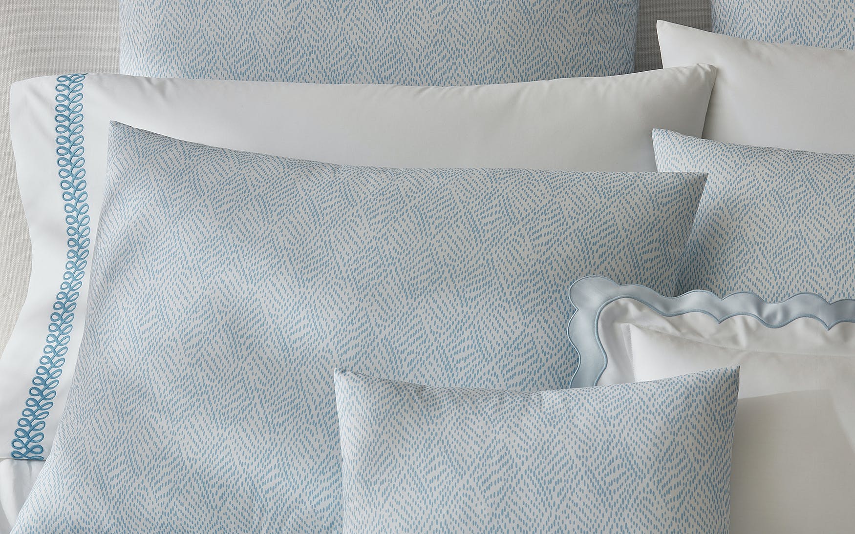 A close-up view of neatly arranged pillows and pillowcases from the Duma Diamond Bedding Collection by Matouk, showcasing light blue and white patterns in luxurious Egyptian cotton percale with both solid and decorative trim designs, accented by subtle geometric motifs.