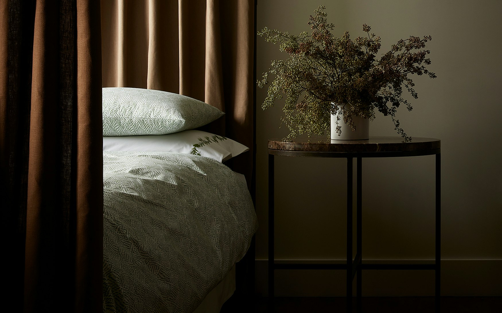 A neatly made bed is dressed in Matouk’s Duma Diamond Bedding Collection, crafted from luxurious Egyptian cotton percale, standing beside a round table with a vase of dried flowers and framed by brown curtains in the background.