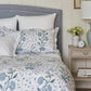 A bed is made up with Matouk's Schumacher Khilana Bedding Collection in blue, featuring a blue and white floral comforter, white pillows, and a gray headboard beside a nightstand topped with a lamp, clock, flowers, and stacked books.