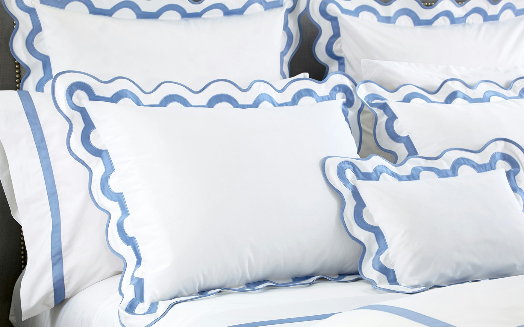 White pillows and bedding with blue scalloped trim from the Matouk Mirasol Bedding Collection are neatly arranged on a bed, crafted from luxurious 600 thread count Egyptian cotton percale.
