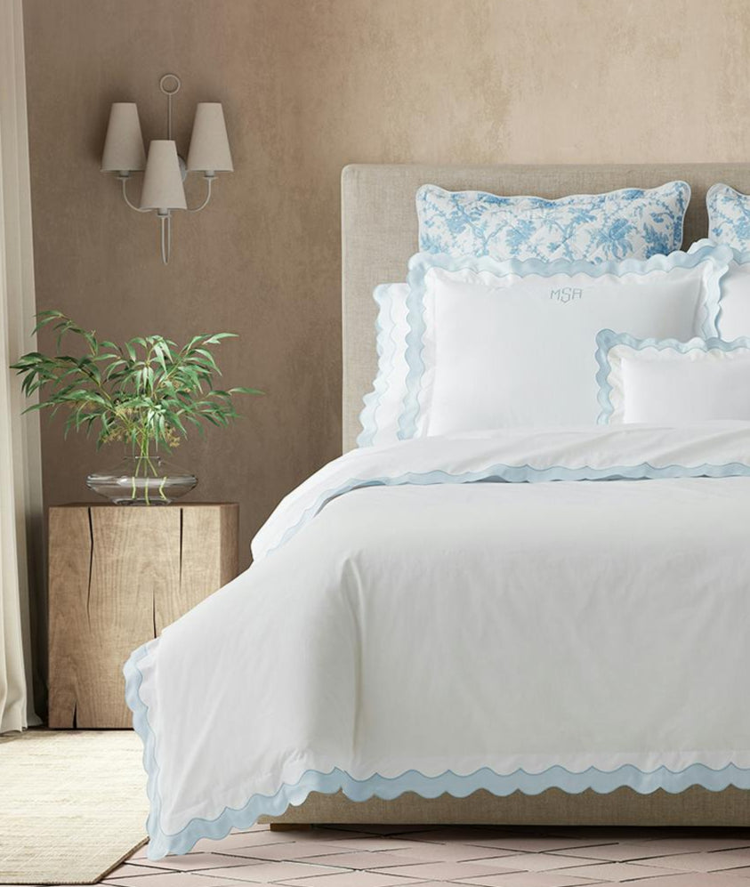 A bed dressed in Matouk’s Lorelei Bedding Collection, Blue—OEKO-TEX® certified, 600 thread count white linens with light blue scalloped edges—paired with decorative pillows and a wooden nightstand in a serene bedroom.