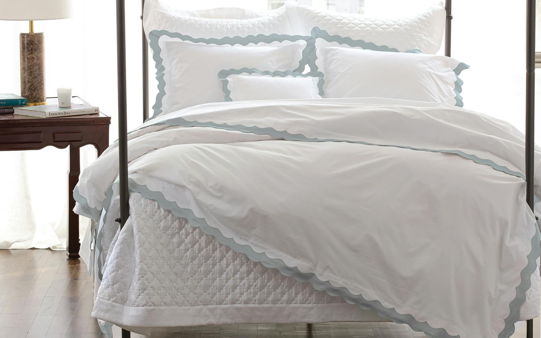 The Matouk Lorelei Bedding Collection in Pool, featuring a white bed with light blue scallop trim on pillows and duvet by Matouk, complements a wooden side table adorned with books, a lamp, and a candle for an artisanal look.