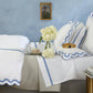 A neatly made bed with Matouk’s Alba Bedding Collection in Silver—OEKO-TEX® Standard 100 certified sateen linens, white with blue trim, decorative pillows, a crown and teacup on the bed, books stacked beside it, plus a bouquet of white flowers.