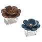 Two Kim Seybert Begonia Napkin Rings, one in a rich brown reminiscent of faux wood and the other in a striking blue—each adorned with metallic accents—are elegantly displayed on clear rectangular bases against a white background.