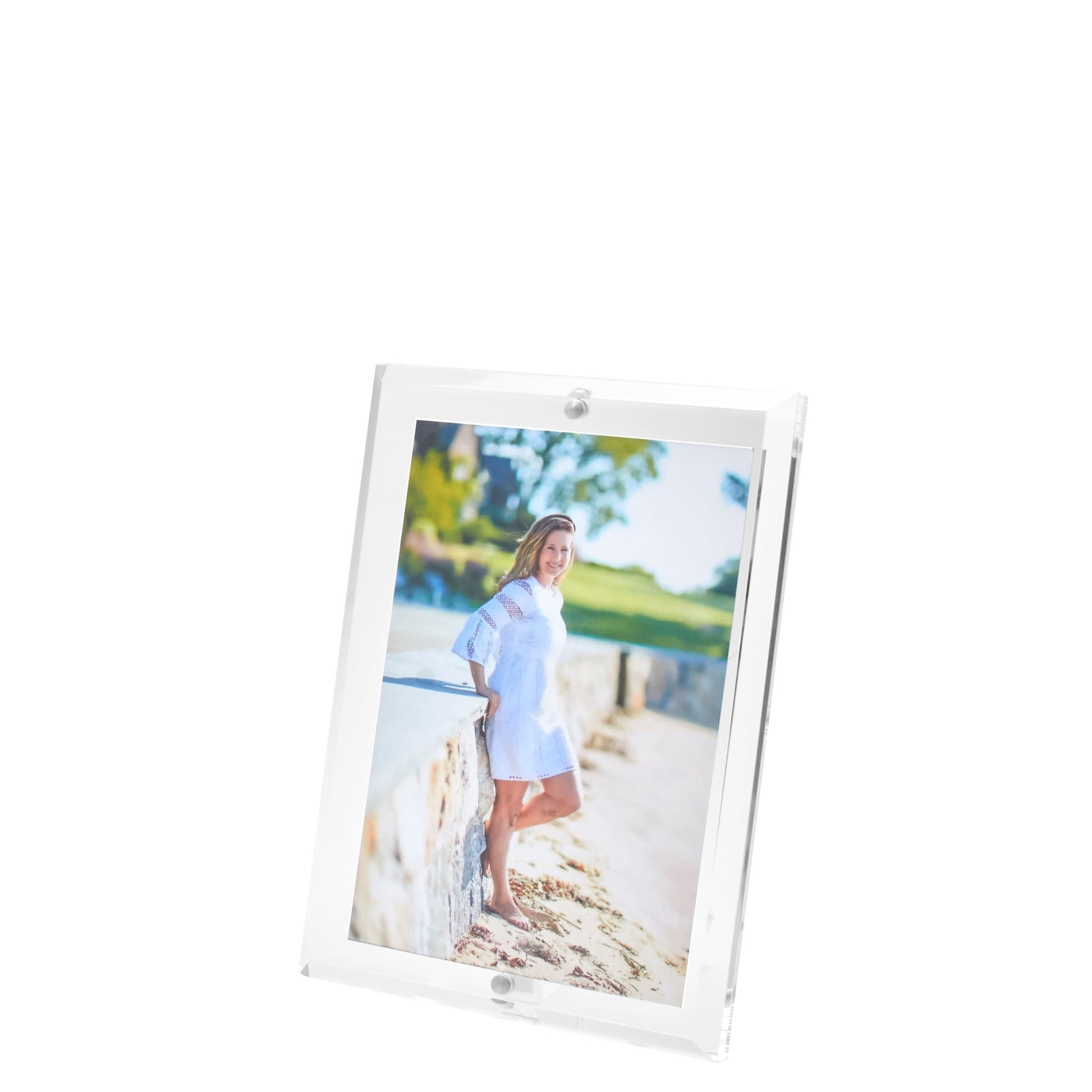 The Clear Beveled Frame Collection by Tara Wilson Designs showcases a portrait of a woman in a white dress standing outdoors near a stone wall and greenery.