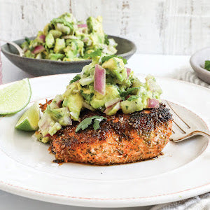 A seasoned salmon fillet topped with avocado salsa sits on a white plate with a lime wedge, perfectly capturing the essence of Simon & Schuster’s Coastal Kitchen. In the background, a fork and bowl of salsa complete this fresh take on healthy seafood meals.