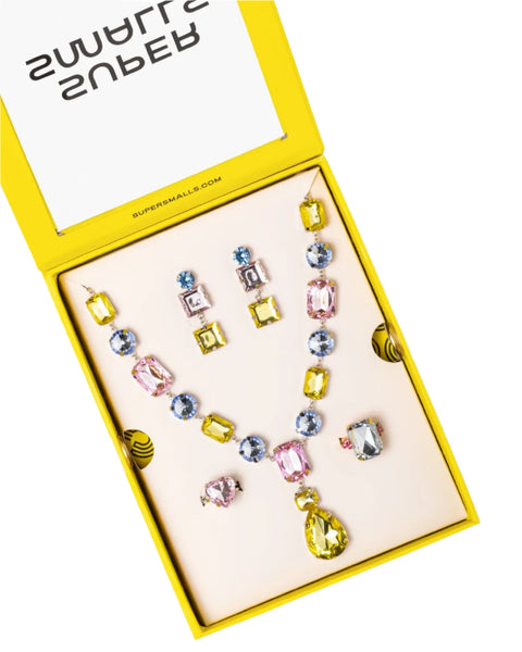 The Super Smalls Black Tie Mega Set comes in a cheerful yellow box and includes a necklace adorned with supersized gemstones in various vibrant colors, matching earrings, and an adjustable ring. The top of the box features mirrored text that reads "Supersmalls.com".