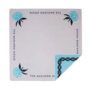 The Mahjong House Double-Sided Travel Mahjong Mats feature blue peony designs and “THE MAHJONG HOUSE” text on each edge, with a folded corner revealing a blue floral pattern—perfect as a portable mahjong mat for travel.
