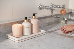 A marble bathroom countertop displays L'Avant Collective's Blushed Bergamot Candle and two soap dispensers on a tray beside a sink, filling the space with a luxurious scent from its soy and coconut blend.