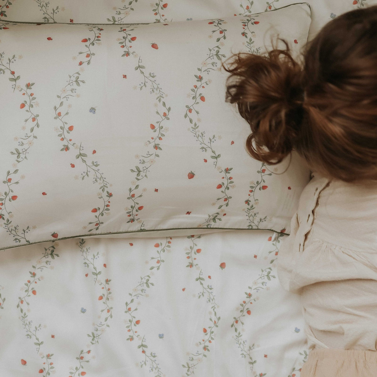 A person with brown hair lies on a bed adorned with the Botanical Berries Bedding Collection by Gooselings, featuring a white pillow and sheet patterned with delicate strawberries and vines. The cozy scene evokes bedtime dreams, while the charming bedding recalls classic children’s collections. The person’s face is not visible.