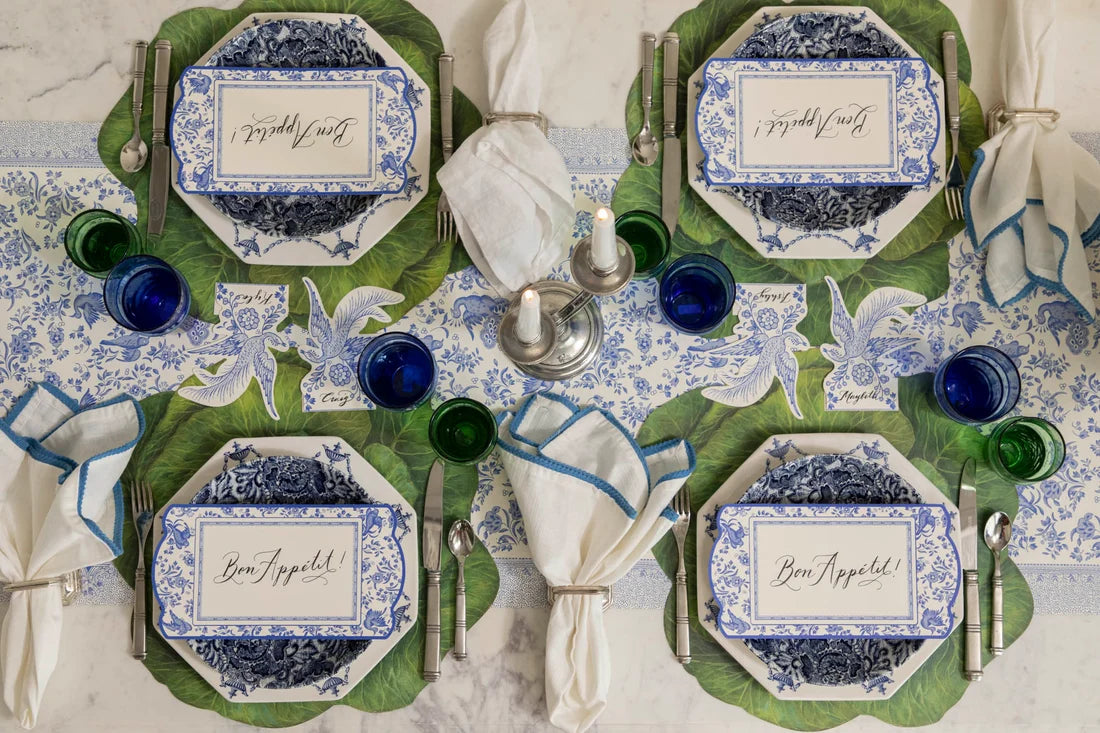 A table set for four with blue and white plates, green glasses, silver-ringed white napkins, Hester & Cook Die Cut Cabbage Placemats, place cards that say “Bon Appetit!”, all arranged on a floral table runner.