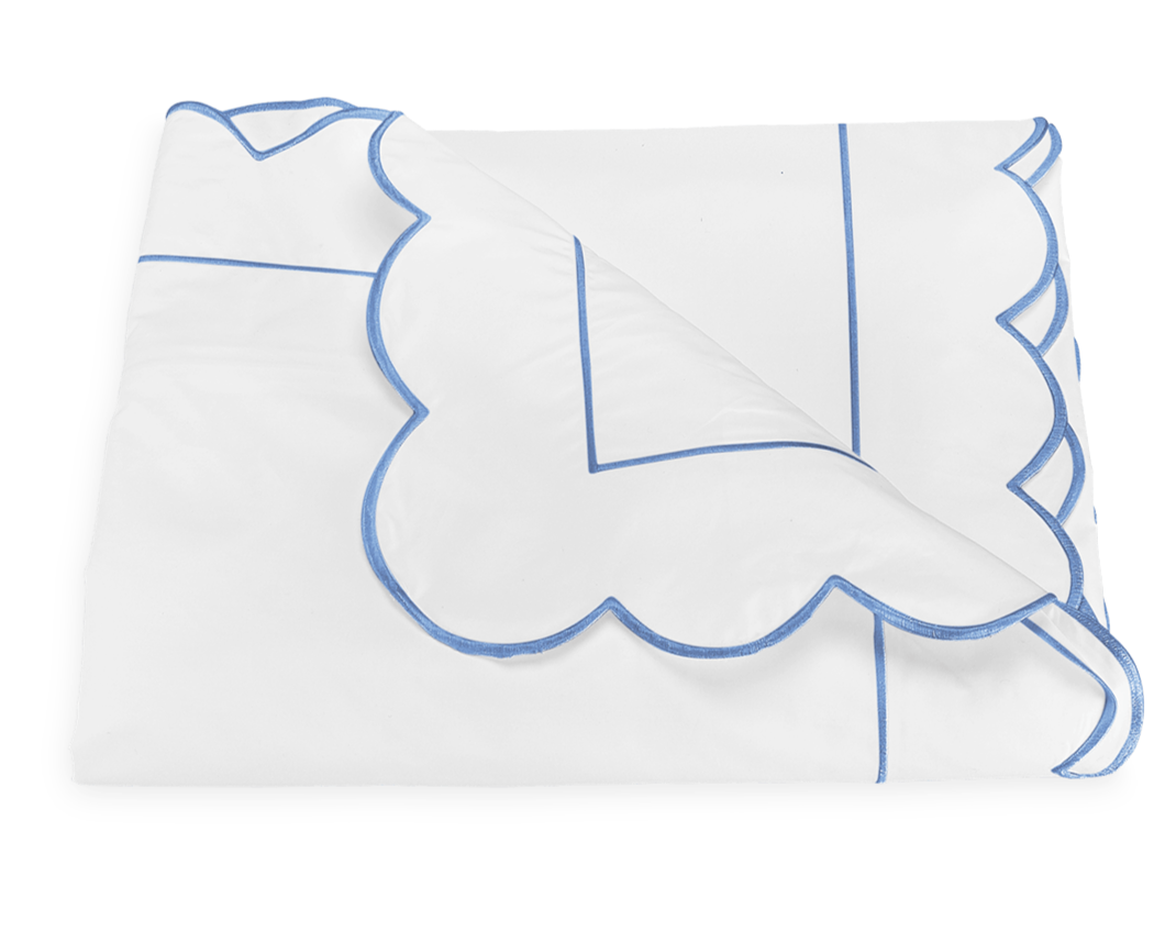 A folded white blanket from the Matouk Butterfield Bedding Collection, Ocean, featuring scalloped edges with blue trim is displayed on a plain background.