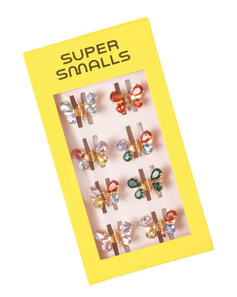 A yellow box from the brand "Super Smalls," labeled "Super Smalls Talent Show Butterfly Hair Clips," contains six pairs of butterfly-shaped earrings adorned with multicolored gemstones, each pair fastened to the box with small gray clips.