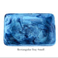 A small, rectangular tray from Nashi Home's Denim Resin Serveware Collection is shown with a blue marbled pattern on a white background. Made from shatter-resistant resinware, it's perfect for stylish home decor. Text below: "Rectangular Tray Small.