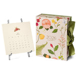 The Karen Adams 2026 Classic Desk Calendar Collection, featuring floral designs and a green ribbon, is displayed with an August 2025 card on a small metal easel—an ideal gift and a best-selling choice.