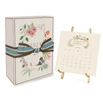 A 2027 Classic Desk Calendar box tied with a striped ribbon next to an open January 2027 calendar displayed on a gold stand.