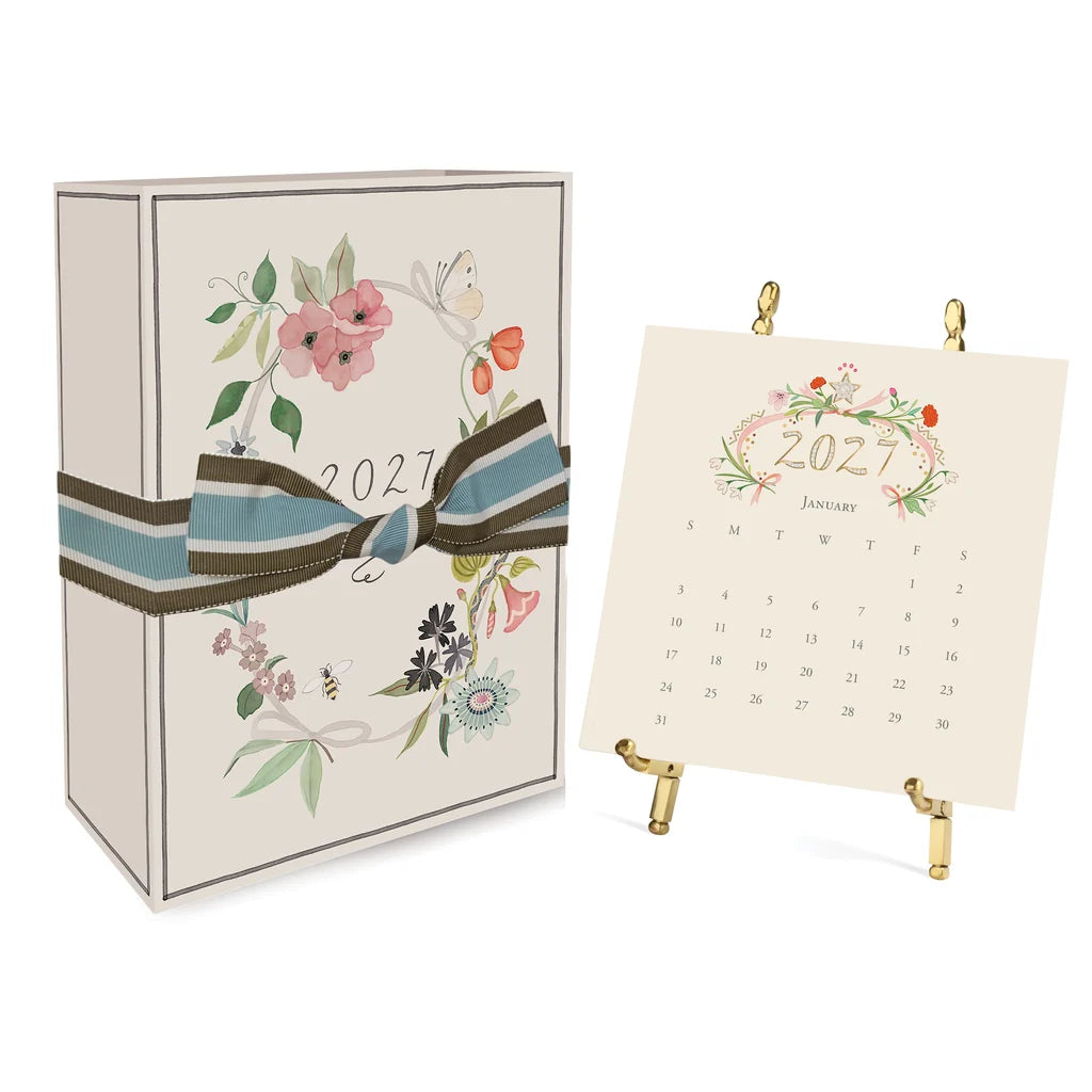 A 2027 Classic Desk Calendar box tied with a striped ribbon next to an open January 2027 calendar displayed on a gold stand.