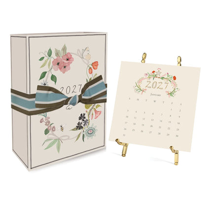 A 2027 Classic Desk Calendar box tied with a striped ribbon next to an open January 2027 calendar displayed on a gold stand.