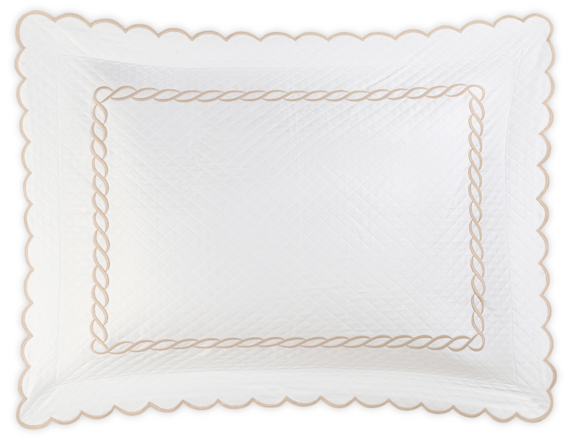 The Matouk Classic Chain Scallop Matelassé Bedding Collection in Dune features a white quilted pillow sham with scalloped edges and a beige chain-inspired border, shown against a black background.