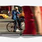 An open copy of Assouline’s "5th Avenue" displays an iconic street photo of a man taking a picture beside a bicycle, with a blurred figure and yellow taxis in the background.