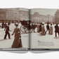 An open history book from Assouline titled "5th Avenue" shows a sepia photo of people in late 19th-century attire walking by a horse-drawn carriage, with historic buildings lining the iconic New York street.