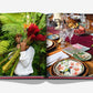 The open book, "Santo Domingo" by Assouline, reveals one page with a person amid green leaves and red flowers, and another showcasing a table setting with plates, glasses, fruits, and nuts—capturing the vibrant culture of Santo Domingo's Zona Colonial.