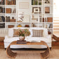 A white sofa with plaid pillows faces built-in bookshelves; in front, the Selamat Healdsburg Wicker Coffee Table displays books and a vase of dried flowers. Stairs are visible to the right.