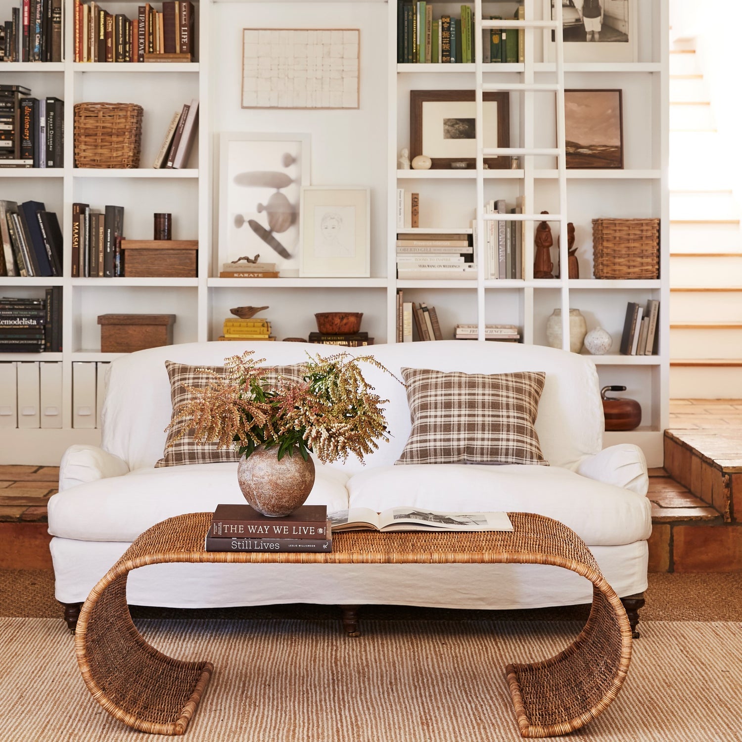 A white sofa with plaid pillows faces built-in bookshelves; in front, the Selamat Healdsburg Wicker Coffee Table displays books and a vase of dried flowers. Stairs are visible to the right.