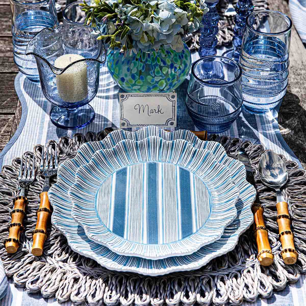 A table setting featuring Juliska’s Cabana Stripe Melamine Dinnerware Collection, bamboo-handled cutlery, blue glassware, a candle, a floral centerpiece, and a place card labeled "Mark.