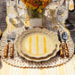 A place setting with Juliska's Cabana Stripe Melamine Dinnerware Collection—yellow striped plates with ruffle trim, bamboo-handled cutlery, and clear glasses on a braided placemat. A bouquet of yellow flowers and a name card appear in the background.