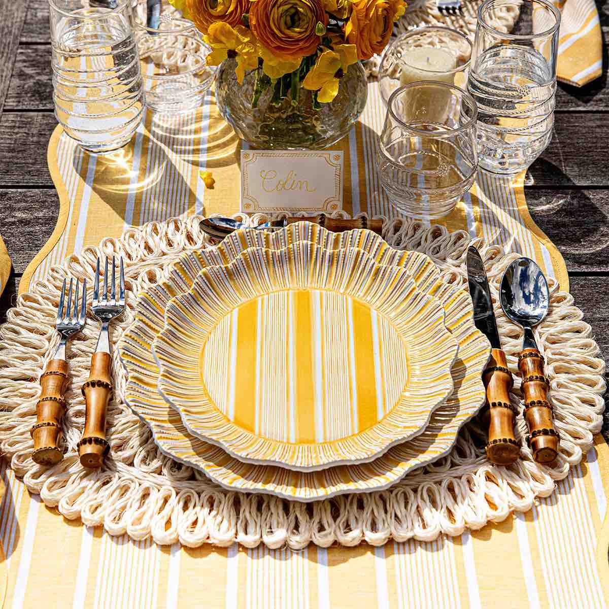 A place setting with Juliska's Cabana Stripe Melamine Dinnerware Collection—yellow striped plates with ruffle trim, bamboo-handled cutlery, and clear glasses on a braided placemat. A bouquet of yellow flowers and a name card appear in the background.